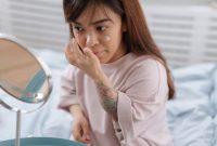 Seorang perempuan sedang mengaplikasikan pelembap pada wajahnya yang kering, menunjukkan pentingnya langkah hidrasi dalam rutinitas perawatan kulit. Pelembap yang tepat membantu mengunci kelembapan dan memperbaiki *skin barrier* kulit kering.