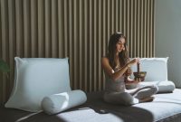 Seseorang sedang menikmati secangkir teh herbal sambil melihat pemandangan kota dari jendela, menunjukkan momen ketenangan dan mindfulness di tengah kesibukan perkotaan. Gambar ini merefleksikan pentingnya menciptakan ruang untuk relaksasi dalam gaya hidup sehat.