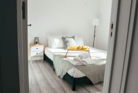 Sebuah kamar tidur minimalis dengan dominasi warna putih dan abu-abu muda, dilengkapi furnitur kayu sederhana dan pot tanaman hijau, menciptakan suasana yang menenangkan dan rapi. Desain interior ini menonjolkan prinsip 