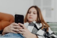 Seorang remaja yang tampak murung sedang menatap layar ponselnya, merefleksikan isolasi dan tekanan yang seringkali dialami generasi muda di tengah arus deras dunia digital. Gambar ini menggambarkan pentingnya perhatian dan dukungan terhadap kesehatan mental mereka di era modern ini.