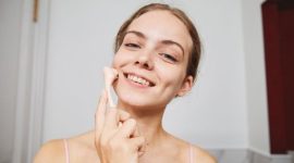 Seorang remaja perempuan sedang tersenyum sambil memegang produk perawatan kulit di kamar mandi, menunjukkan pentingnya rutinitas skincare yang tepat untuk kulit muda. Pemilihan produk yang lembut dan sesuai kebutuhan kulit remaja sangat penting untuk menjaga kesehatan dan kecerahan alami kulit mereka.