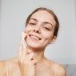 Seorang remaja perempuan sedang tersenyum sambil memegang produk perawatan kulit di kamar mandi, menunjukkan pentingnya rutinitas skincare yang tepat untuk kulit muda. Pemilihan produk yang lembut dan sesuai kebutuhan kulit remaja sangat penting untuk menjaga kesehatan dan kecerahan alami kulit mereka.