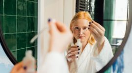 Seorang wanita tersenyum sambil mengaplikasikan serum wajah di pagi hari, menggambarkan rutinitas perawatan kulit yang konsisten untuk mendapatkan kulit sehat dan bercahaya. Rutinitas perawatan wajah yang teratur merupakan investasi jangka panjang untuk kesehatan kulit optimal.