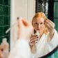Seorang wanita tersenyum sambil mengaplikasikan serum wajah di pagi hari, menggambarkan rutinitas perawatan kulit yang konsisten untuk mendapatkan kulit sehat dan bercahaya. Rutinitas perawatan wajah yang teratur merupakan investasi jangka panjang untuk kesehatan kulit optimal.
