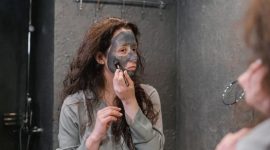 Seorang wanita tersenyum sembari mengaplikasikan masker wajah alami dari bahan-bahan dapur, menunjukkan kemudahan dan kesenangan dalam merawat kulit secara tradisional di rumah.