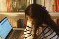 Seorang mahasiswa terlihat kewalahan dengan tumpukan buku dan tugas di depan laptopnya di kampus, menggambarkan tekanan akademik yang sering dialami selama masa perkuliahan. Ekspresi kelelahan dan cemas terlihat jelas pada wajahnya, merefleksikan pentingnya strategi pengelolaan stres.