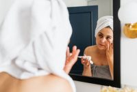 Seorang wanita sedang mengaplikasikan pelembap pada wajahnya yang kering, menunjukkan momen penting dalam rutinitas perawatan untuk mengembalikan hidrasi dan elastisitas kulit. Tindakan sederhana ini merupakan kunci untuk mendapatkan kulit wajah yang sehat dan terhindar dari sensasi tertarik akibat kering.