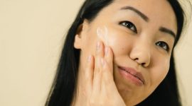 Seorang wanita sedang mengaplikasikan pelembap wajah, menunjukkan pentingnya hidrasi dalam rutinitas perawatan kulit sehari-hari untuk menjaga elastisitas dan cahaya alami kulit.